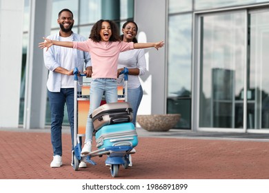 Family Trip Concept. Portrait of cheerful African American girl having fun and spreading hands, ready for vacation, standing on luggage cart. Parents walking with baggage trolley, riding daughter