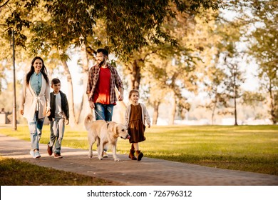 Family With Two Children Walking Down The Road In Autumn Park With A Dog