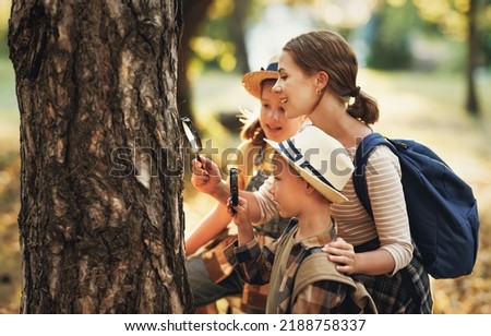 Нарру family: two   kids boy and girl  with backpacks looking examining tree bark through magnifying glass while exploring forest nature and environment   during outdoor ecology school lesson