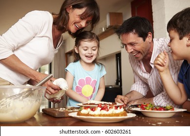 A Family, Two Parents And Two Children In The Kitchen Icing A Cake With Fruit And Cream