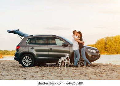 Family Of Two Person And Dog Embrace Near Car On Sunset River