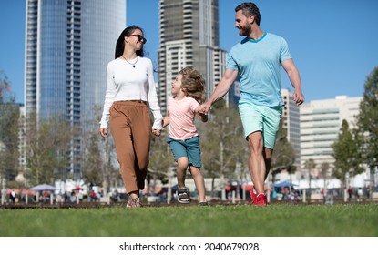 Family In Urban Skyline On Park Lawn. Happy Parents Holding Hands With Son And Walking In Sunny Summer Citi Street, Funny Family Moment.