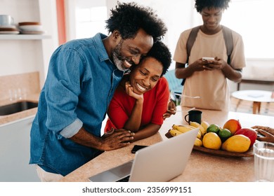 Family using technology in the morning, with parents engaging in a video call on a laptop while their teenage son uses his phone in the background.  Modern family using mobile devices in the kitchen.  - Powered by Shutterstock