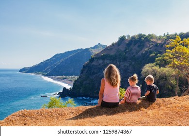 Family Vacation Lifestyle. Happy Mother, Kids On Hill With Scenic View Of High Cliffs, Fishers Village On Black Beach. Children Looking At Blue Sea. Bukit Asah Is Popular Travel Destination In Bali.