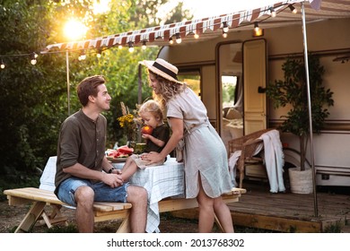 Family vacation in mobile home: young parents travel with small preschool son have picnic on terrace on sunset at rv camper trailer. People enjoy summer road voyage on caravan car. Summer outdoor trip