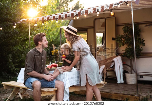 Family vacation in mobile home: young parents travel\
with small preschool son have picnic on terrace on sunset at rv\
camper trailer. People enjoy summer road voyage on caravan car.\
Summer outdoor trip