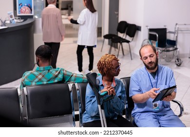 Family Waiting Room At Hospital Rehabilitation Floor. Health Professional Showing File To Laughing Hair Girl Using Crutches. Young African American Man Waiting Seated In Lobby Chairs Of Medical Tower.