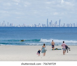 Family Walk Across Beach. Scenic Beach Scene With Parents With Child And Surfer. Family Beach Holiday - Rainbow Bay, Gold Coast, Australia With Surfers Paradise Skyline In Background.