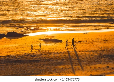 Family Walking Together On The Beach At The Sunset. Group Of People With Pet Walks On The Seashore At The Golden Hour During Summer Holiday. Mother, Father And Sons With Dog On The Beach At Sunrise.