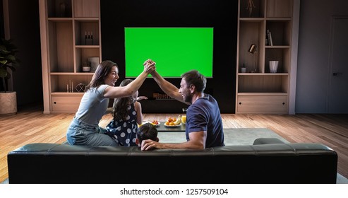 A Family Is Watching A TV And Celebrating Some Joyful Moment, Sitting On The Couch In The Living Room. The Living Room Is Made In 3D. TV Is Green Screen For Further Editing.