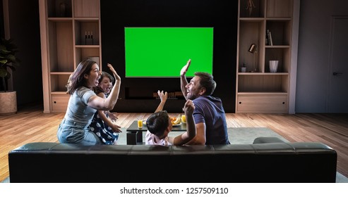 A Family Is Watching A TV And Celebrating Some Joyful Moment, Sitting On The Couch In The Living Room. The Living Room Is Made In 3D. TV Is Green Screen For Further Editing.