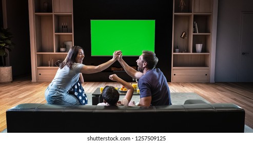 A Family Is Watching A TV And Celebrating Some Joyful Moment, Sitting On The Couch In The Living Room. The Living Room Is Made In 3D. TV Is Green Screen For Further Editing.