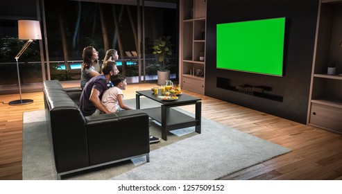 A Family Is Watching A TV And Celebrating Some Joyful Moment, Sitting On The Couch In The Living Room. The Living Room Is Made In 3D. TV Is Green Screen For Further Editing.