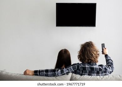 Family Watching TV Sitting On Couch At Home, Rear View. Couple Enjoying Movie Switching Channel Pointing Remote Control At Black Flatscreen Plasma Set In Living Room Indoors. Selective Focus