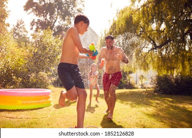 Family Wearing Swimming Costumes Having Water Fight With Water Pistols In Summer Garden