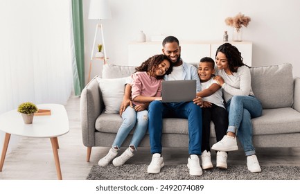 Family Weekend. Portrair of cheerful African American parents and their little children using laptop at home together, watching movie or browsing internet, sitting on the couch in living room - Powered by Shutterstock