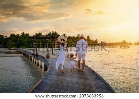 A family in white summer clothes walks holding hands over a wooden pier towards a tropical island in the Maldives during sunset time