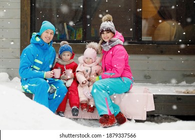 Family In Winter Clothes Is Sitting On The Porch And Drinking Tea Or Coffee. Country House In A Snowy Coniferous Forest. Warm Jackets, Knitted Hats, Cups. Family Happiness, Values And Traditions.