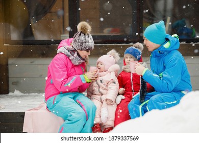 Family In Winter Clothes Is Sitting On The Porch And Drinking Tea Or Coffee. Country House In A Snowy Coniferous Forest. Warm Jackets, Knitted Hats, Cups. Family Happiness, Values And Traditions.