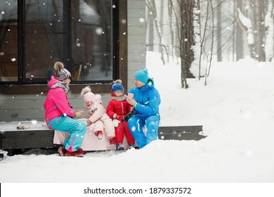 Family In Winter Clothes Is Sitting On The Porch And Drinking Tea Or Coffee. Country House In A Snowy Coniferous Forest. Warm Jackets, Knitted Hats, Cups. Family Happiness, Values And Traditions.