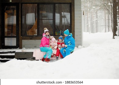 Family In Winter Clothes Is Sitting On The Porch And Drinking Tea Or Coffee. Country House In A Snowy Coniferous Forest. Warm Jackets, Knitted Hats, Cups. Family Happiness, Values And Traditions.