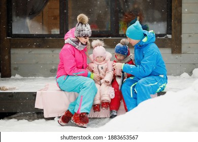 Family In Winter Clothes Is Sitting On The Porch And Drinking Tea Or Coffee. Country House In A Snowy Coniferous Forest. Warm Jackets, Knitted Hats, Cups. Family Happiness, Values And Traditions.