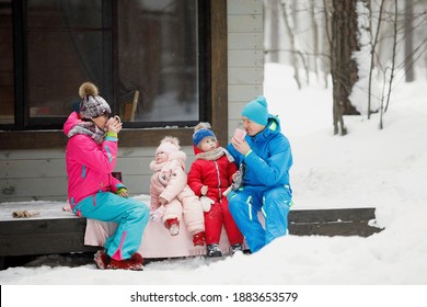 Family In Winter Clothes Is Sitting On The Porch And Drinking Tea Or Coffee. Country House In A Snowy Coniferous Forest. Warm Jackets, Knitted Hats, Cups. Family Happiness, Values And Traditions.