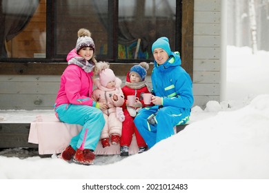 Family In Winter Clothes Is Sitting On The Porch And Drinking Tea Or Coffee. Country House In A Snowy Coniferous Forest. Warm Jackets, Knitted Hats, Cups. Family Happiness, Values And Traditions.