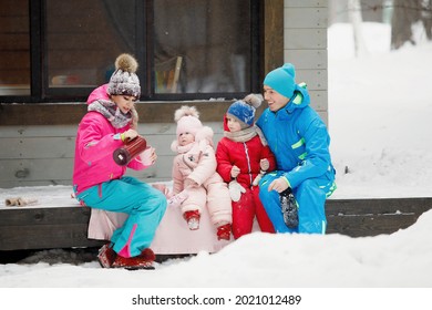 Family In Winter Clothes Is Sitting On The Porch And Drinking Tea Or Coffee. Country House In A Snowy Coniferous Forest. Warm Jackets, Knitted Hats, Cups. Family Happiness, Values And Traditions.