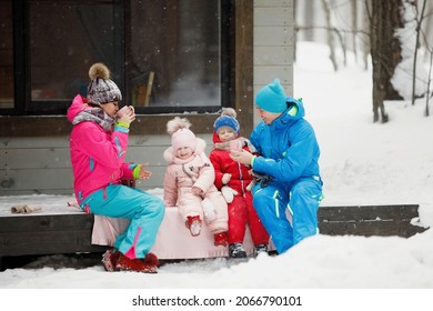 Family In Winter Clothes Is Sitting On The Porch And Drinking Tea Or Coffee. Country House In A Snowy Coniferous Forest. Warm Jackets, Knitted Hats, Cups. Family Happiness, Values And Traditions.