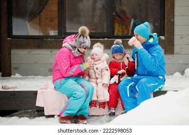 Family In Winter Clothes Is Sitting On The Porch And Drinking Tea Or Coffee. Country House In A Snowy Coniferous Forest. Warm Jackets, Knitted Hats, Cups. Family Happiness, Values And Traditions.