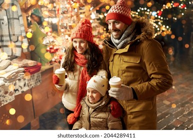 Family, Winter Holidays And Celebration Concept - Happy Mother, Father And Little Daughter With Takeaway Drinks At Christmas Market On Town Hall Square In Tallinn, Estonia Over Lights