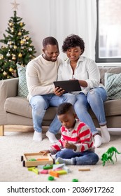 Family, Winter Holidays And People Concept - Happy African American Mother And Father Using Tablet Pc Computer And Baby Son Playing With Toy Blocks And Dinosaur At Home On Christmas