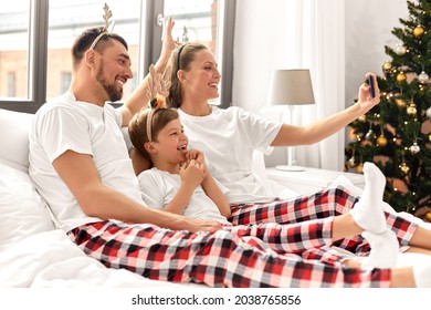 Family, Winter Holidays And People Concept - Happy Mother, Father And Little Son In Matching Pajamas Taking Selfie With Smartphone In Bed In Christmas Morning