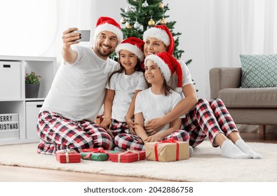 Family, Winter Holidays And People Concept - Happy Mother, Father And Two Daughters In Santa Hats Sitting Under Christmas Tree At Home And Taking Selfie With Smartphone