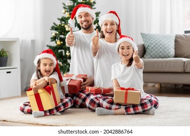 Family, Winter Holidays And People Concept - Happy Mother, Father And Two Daughters In Matching Pajamas Sitting With Gifts Under Christmas Tree At Home Showing Thumbs Up