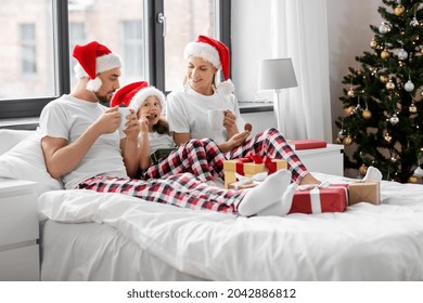 Family, Winter Holidays And People Concept - Happy Mother, Father And Little Daughter With Christmas Gifts Drinking Coffee And Eating Coookies In Bed At Home