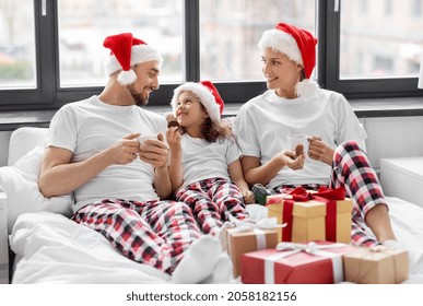 Family, Winter Holidays And People Concept - Happy Mother, Father And Little Daughter With Christmas Gifts Drinking Coffee And Eating Coookies In Bed At Home