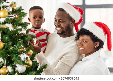 Family, Winter Holidays And People Concept - Happy African American Mother, Father And Baby Son Decorating Christmas Tree At Home On