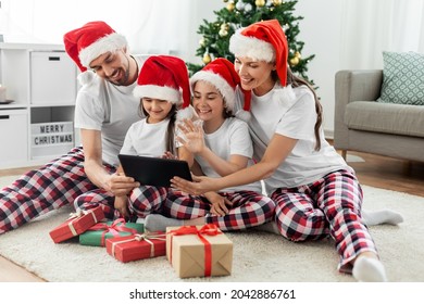 Family, Winter Holidays And Technology Concept - Happy Mother, Father And Two Daughters In Santa Hats With Christmas Gifts Sitting On Floor And Having Video Call On Tablet Pc Computer At Home