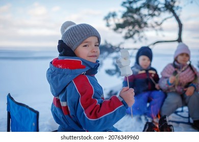 Family Winter Picnic. Happy Friends Sit Around Campfire On The Shore Of Frozen Lake In Forest And Fry Marshmallows During A Winter Trip At Weekend