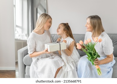 Family Woman - Grandmother, Daughter-mother, Granddaughter - Give Each Other Flowers And Gifts For The Holiday On 8-March. Beautiful Women And A Little Girl In White Clothes At Home On The Couch.