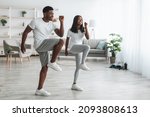 Family Workout. African American Husband And Wife Training Together In Living Room, Doing High Knees Exercise. Happy Black Couple Warming Up, Standing And Lifting Leg Up To Chest, Free Copy Space