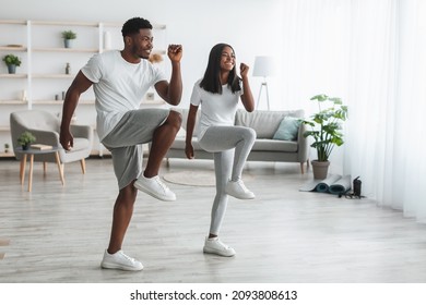 Family Workout. African American Husband And Wife Training Together In Living Room, Doing High Knees Exercise. Happy Black Couple Warming Up, Standing And Lifting Leg Up To Chest, Free Copy Space