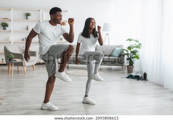 Family Workout. African American Husband And Wife\
Training Together In Living Room, Doing High Knees Exercise. Happy\
Black Couple Warming Up, Standing And Lifting Leg Up To Chest, Free\
Copy Space