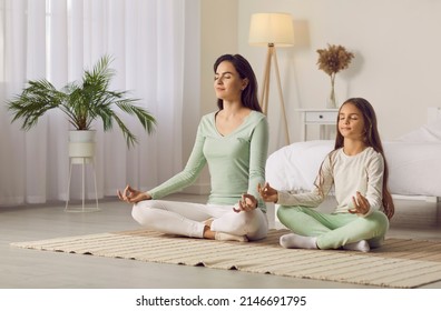 Family yoga. Beautiful young woman and her charming little daughter are smiling while doing yoga together at home. Family sits in lotus position on floor in living room. Mom teaches child to meditate. - Powered by Shutterstock