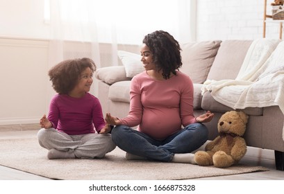 Family yoga concept. Pregnant black mother and her cute little daughter meditating together, sitting on floor in lotus position, free space