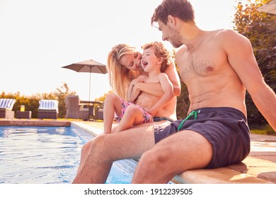 Family With Young Son Sitting On Edge Of Pool Having Fun On Summer Vacation In Outdoor Swimming Pool