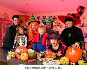 Family/friends Posing For The Camera In Their Halloween Costumes. They Are Standing In The Kitchen With Party Food And Treats Set Out In Front Of Them. 