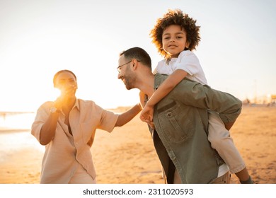 Family's beach adventure. Young parents walking with son on seaside, father piggybacking boy outdoors, enjoying free time on nature at sunset - Shutterstock ID 2311020593